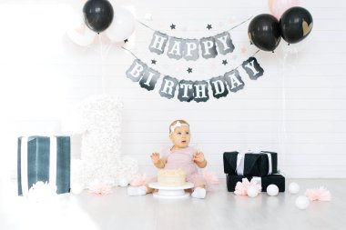 Bir yaşında bir kız ilk doğum günü şerefine tatlı bir pastanın yanında yerde oturuyor.