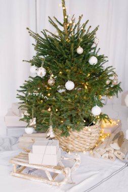 Beyaz oyuncaklarla süslenmiş hasır bir tencerede Noel süslenmiş ağaç. Ağacın yanında hediyeli ahşap kızaklar var.