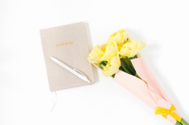 İş yerinde bej bir günlük, beyaz bir kalem, beyaz arka planda bir buket sarı lale. Daire yatıyordu, yukarıdan görünüyordu. Düz ofis masası.