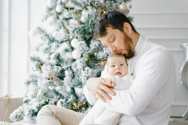 Genç bir baba genç oğlunu öper ve parlak oturma odasındaki Noel ağacının yanında oturur.