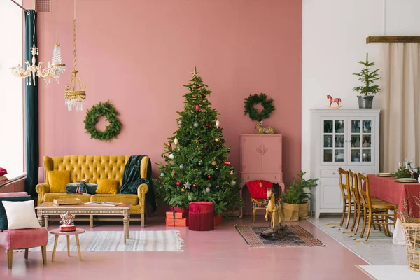 Noel ve Yeni Yıl için dekore edilmiş klasik oturma odası: oyuncaklarla süslenmiş Noel ağacı, sarı kanepe, sandalyeli ve dekorlu masa. Pembe oda duvarları