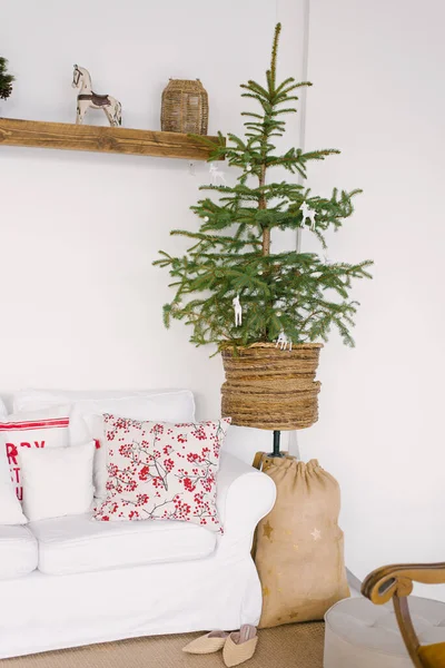 Christmas decor of the living room in the Scandinavian style. White sofa with red pillows, Christmas tree, bag of gifts under it