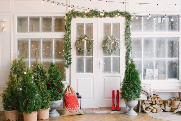 Noel süslerinde ve Noel ağaçlarında beyaz kapısı olan bir veranda. Kapının etrafında çelenkler var. Evin güzel kış terası retro lambalardan çelenklerle dolu.