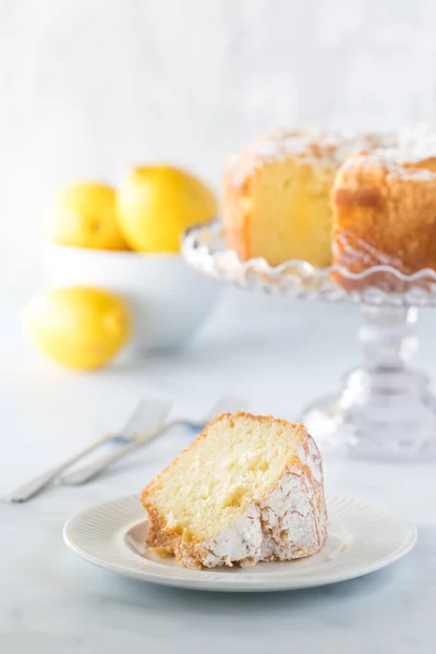 Πρόσοψη μιας φέτας κέικ λεμονιού με το κέικ σε βάση κέικ, πιρούνια και ένα μπολ με λεμόνια σε απαλή εστίαση. — Φωτογραφία Αρχείου