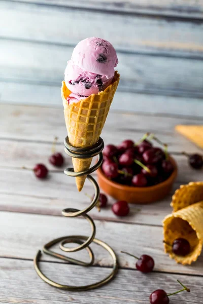 Un seul cône de crème glacée cerise dans un support en spirale métallique avec un bol de cerises derrière . — Photo