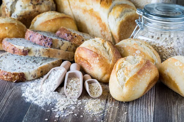 Varios bollos y panes de pan en una mesa de madera con cucharadas de madera de ingredientes utilizados para hacer pan en frente. — Foto de Stock