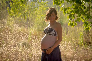 Bir ormanda ayakta, göbeğini gösteren genç bir hamile kadın. O mavi bir etek ve çıplak bir bikini üst giyiyor