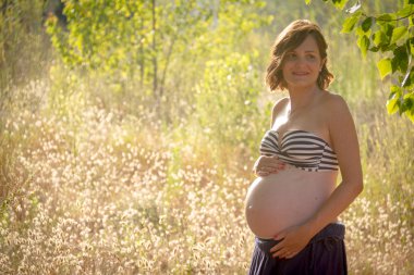 Bir ormanda ayakta, göbeğini gösteren genç bir hamile kadın. O mavi bir etek ve çıplak bir bikini üst giyiyor