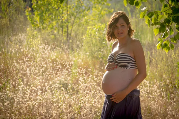 A young pregnant woman showing her belly, standing in a forest. She is wearing a blue skirt and a stripped bikini top Stock Image