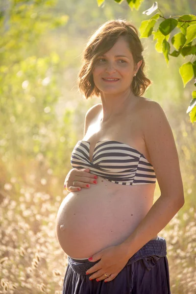 A young pregnant woman showing her belly, standing in a forest. She is wearing a blue skirt and a stripped bikini top Stock Picture