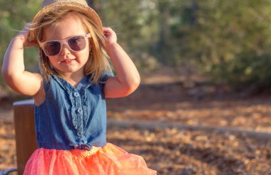 Parkta sevimli sarışın küçük bir kız. Şapka ve güneş gözlüğü takıyor.