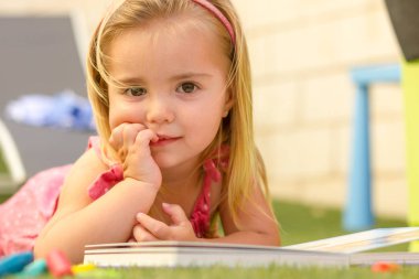 Bir sevimli sarışın küçük kız çim üzerinde döşeme ve bir kitap okuma