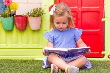 Mavi elbiseli ve kitap okuyan sevimli sarışın küçük bir kız. Çimlerin üzerinde oturuyor ve arka planda renkli bir oyuncak ev var..