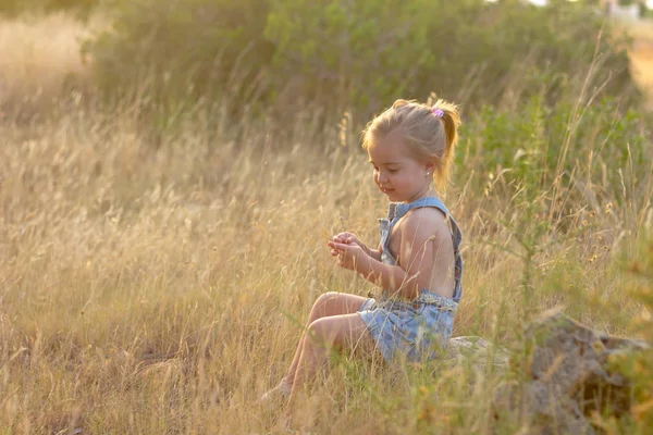 一个可爱的金发小女孩坐在草地上日落。她穿着围兜牛仔裤短裤 — 图库照片