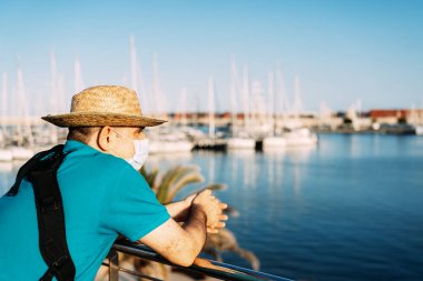 Gün batımında yat limanına bakan yüz maskeli ve şapkalı turist. Akdeniz 'de. Kavram, Coronavirüs zamanlarındaki turist.