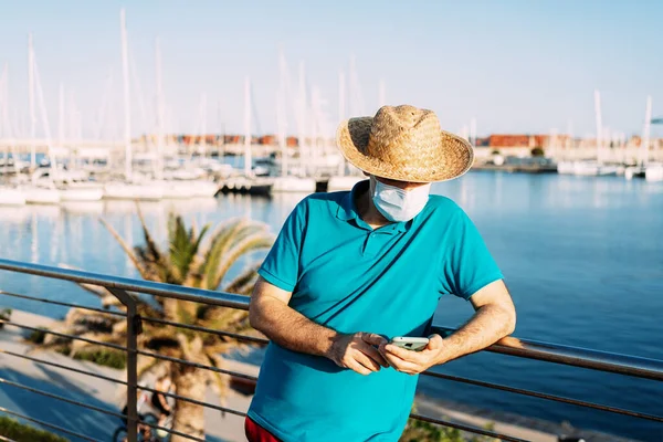Gün batımında marinanın yanında akıllı telefonunun içindekileri kontrol eden maskeli bir turist. Kavram, koronavirüs zamanlarında kişisel koruma.