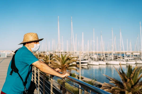 Gün batımında yat limanına bakan yüz maskeli ve şapkalı turist. Akdeniz 'de. Kavram, Coronavirüs zamanlarındaki turist.