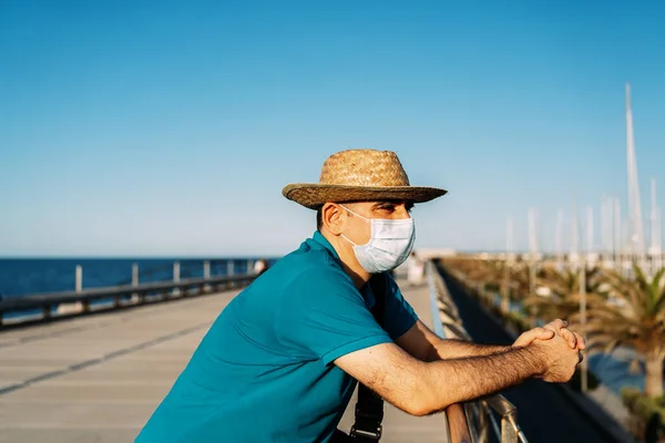 Gün batımında yat limanına bakan yüz maskeli ve şapkalı turist. Akdeniz 'de. Kavram, Coronavirüs zamanlarındaki turist.