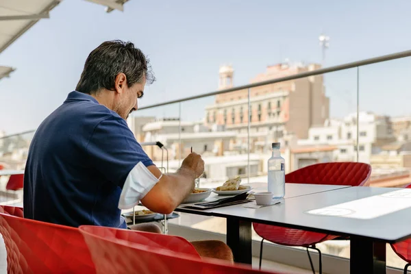 Koluna hijyenik yüz maskesi takmış orta yaşlı bir turist dışarıda manzaralı bir terasta öğle yemeği yiyor..