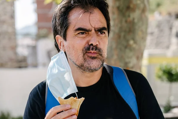 Yetişkin bir erkek kulağından sarkan maskeyle abur cubur yiyor. Yüz maskesi kullanıyor. Yeni normal