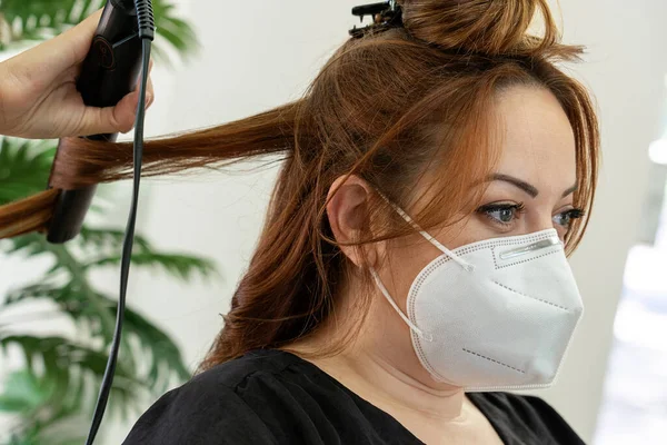 Elegant young hairdresser with face mask straightening the hair of a red hair client\'s in a modern hairdressing salon . Social distancing. New normality