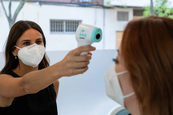 顔マスクテストで保護された若いショップアシスタントは 予防のためのレーザー温度計でインフルエンザをお客様に 新常態 ロイヤリティフリーのストック画像