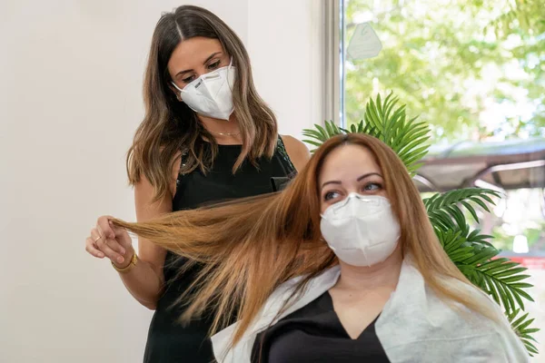 顔のマスクを持つエレガントな若い美容師は 現代の美容室で彼女のクライアントの髪を乾燥させます 社会的距離 ストック写真