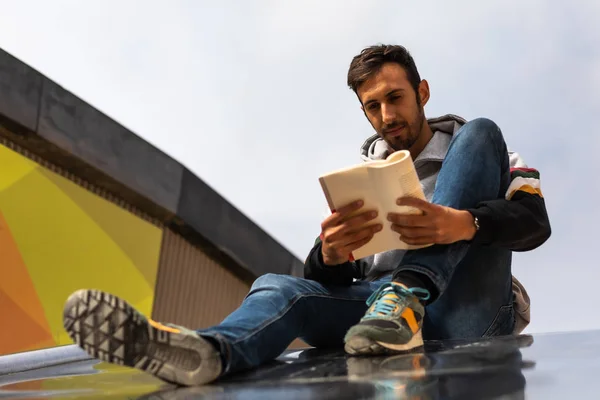 Young Bearded Man Reading A Book Sitting In A Park During The Morning.