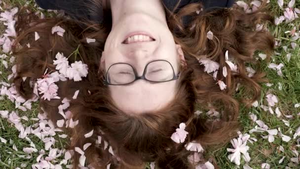 日本の桜の開花を祝う国民の休日に地面に横たわっている若い女性 眼鏡をかけた赤い髪の少女は 花の木から花びらをバラに落とします 高品質4K映像 — ストック動画