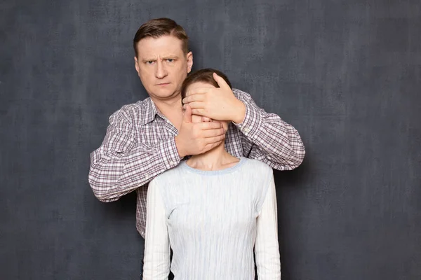 Serious dissatisfied man is covering woman's eyes and mouth