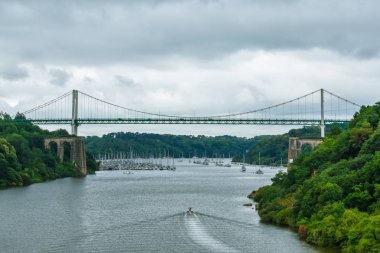 Dramatik bulutlu gökyüzüile yeşil bir doğal ormanda kablo-kaldı köprü. Nehirde motorlu tekne yelken