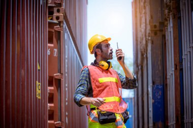 Liman işçisi, liman konteynır nakliye deposunda çalışıyor güvenlik üniforması, şapka, kulaklık takıyor ve telsiz iletişimi kuruyor..