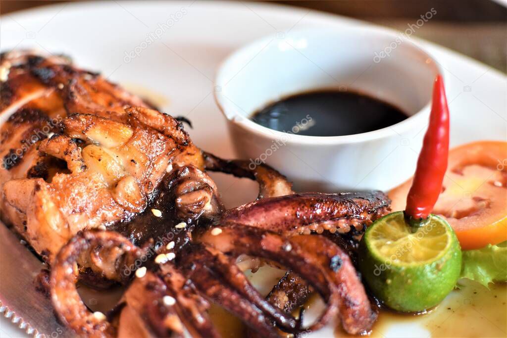 A juicy piece of grilled squid served on a white plate with soy sauce, calamansi, red chili pepper, and a slice of tomato