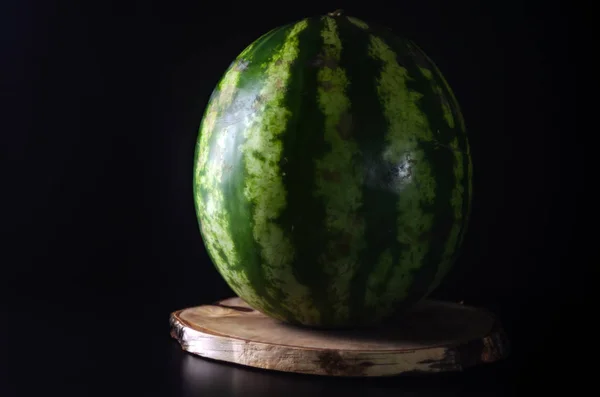 Reife ganze Wassermelone steht auf einem Holzbrett vor schwarzem Hintergrund — Stockfoto