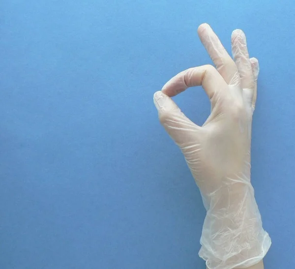 Hand in medical white rubber gloves on a blue background. Okey sign. The concept of healthcare and medical procedures, the prevention of coronavirus or COVID-19. Copy Space.