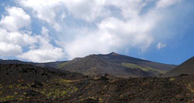 Karanlık volkanik kaya ve toprağın hakim olduğu Etna Dağı 'nın yamaçlarının engebeli ve ıssız manzarası. Doğanın dayanıklılığını gösteren az miktarda yeşil bitki örtüsü. Mavi gökyüzüne karşı heybetli dağ zirvesi.