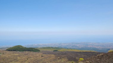 Karanlık volkanik toprak ve bitki örtüsüyle Etna Dağı 'nın engin ve engebeli manzarasının havadan görünüşü. Arka planda kıyı kasabaları ile Akdeniz
