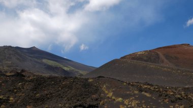 Karanlık volkanik kaya ve toprağın hakim olduğu Etna Dağı 'nın yamaçlarının engebeli ve ıssız manzarası. Doğanın dayanıklılığını gösteren az miktarda yeşil bitki örtüsü. Mavi gökyüzüne karşı heybetli dağ zirvesi.