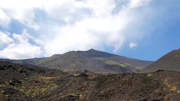 Karanlık volkanik kaya ve toprağın hakim olduğu Etna Dağı 'nın yamaçlarının engebeli ve ıssız manzarası. Doğanın dayanıklılığını gösteren az miktarda yeşil bitki örtüsü. Mavi gökyüzüne karşı heybetli dağ zirvesi.