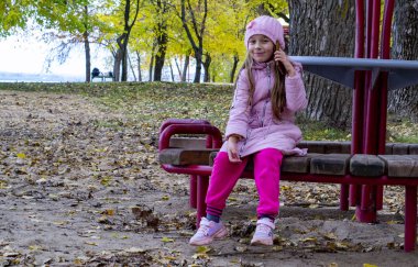 Küçük kız, soğukta sonbahar parktaki bankta oturuyor, ön plana odaklanıyor.