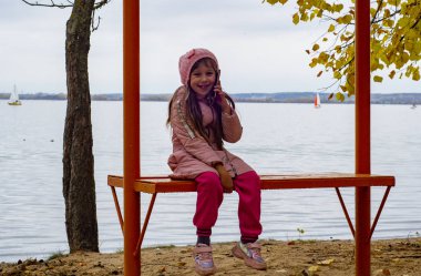 Küçük kız, soğukta sonbahar parktaki bankta oturuyor, ön plana odaklanıyor.