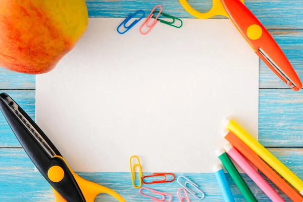 Weißes Papierblatt in der Nähe von Schreibwaren und Apfel auf blauem Holzgrund, Mockup-Konzept für den Schulweg — Stockfoto