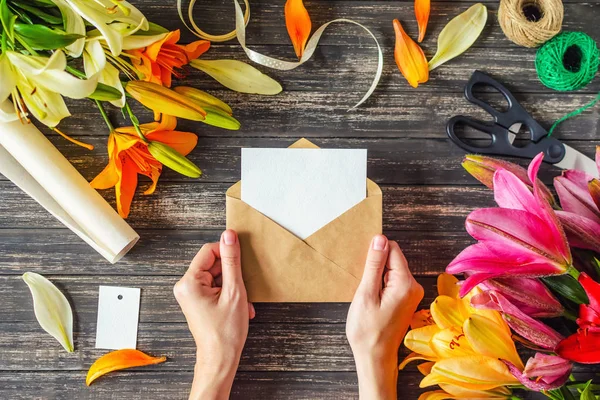 Tarjeta de felicitación de diseño. Las manos de la mujer sostiene sobre con hoja en blanco y decoraciones de flores en la mesa de madera — Foto de Stock