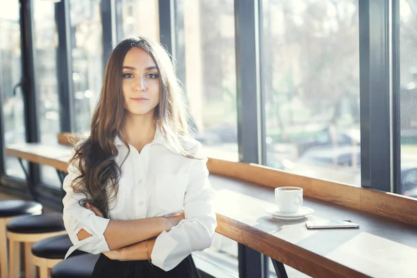 Schöne junge Frau in weißer Bluse trinkt Kaffee und benutzt Smartphone im Café — Stockfoto