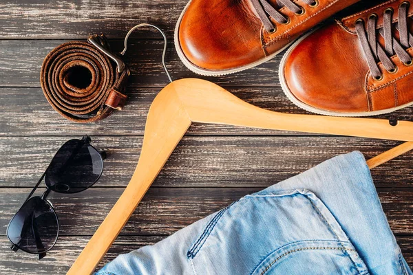 Braune lederne Freizeitschuhe Jeans auf Kleiderbügel, Gürtel und Sonnenbrille auf Holzhintergrund Draufsicht — Stockfoto