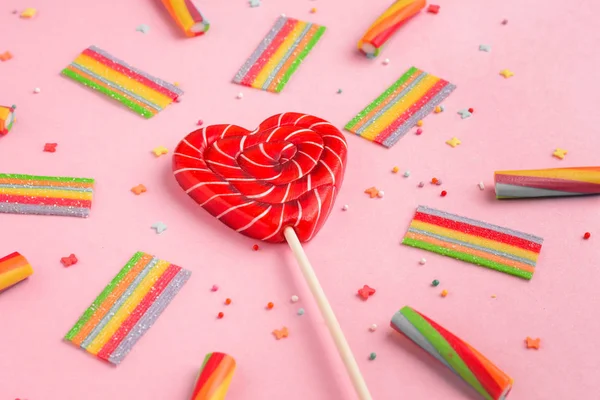 Red lollipop in shape of heart and colorful sweets on a pink background, flat lay