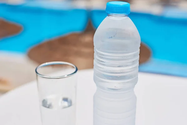 Glas en plastic fles met een water op witte tafel tegen zwembad achtergrond — Stockfoto