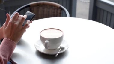 Kafe masasında kahve içen ve akıllı telefon kullanan bir kadın, yavaş çekim videosu. Kız, yemek molasında akıllı telefondan haber akışını kaydırabilir, kapatabilir.