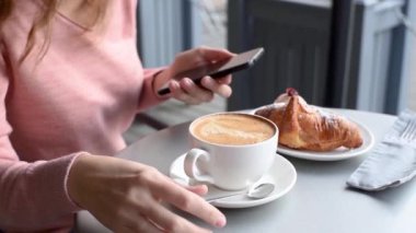 Kafe masasında kahve içen ve akıllı telefon kullanan bir kadın, yavaş çekim videosu. Kız, yemek molasında akıllı telefondan haber akışını kaydırabilir, kapatabilir.