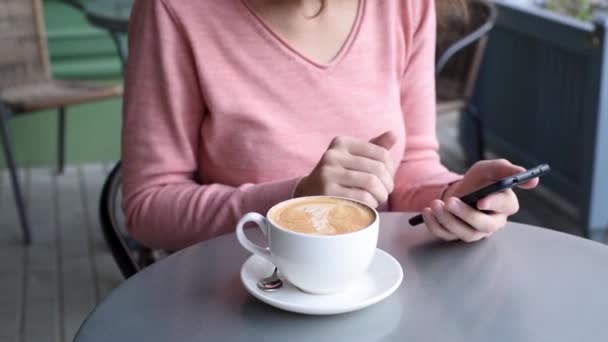 Femme à la table de café en plein air boire du café et en utilisant un smartphone, vidéo au ralenti. Fille fait défiler son flux de nouvelles sur smartphone pendant la pause déjeuner sur la terrasse du restaurant, gros plan 